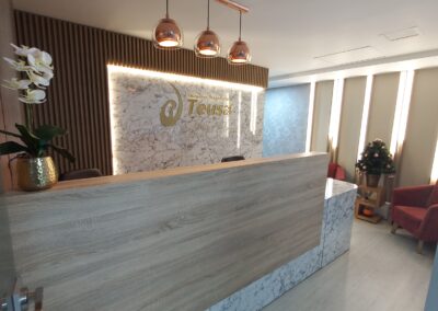 Recepción con acabados en mármol y madera que evocan elegancia, delicadeza y sofisticación. Un logo dorado en acrílico destaca en el centro de un imponente muro de impacto. La iluminación cálida, cuidadosamente integrada, aporta armonía y una sensación acogedora al espacio